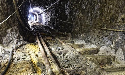 Galleria mineraria oscura, tubi arrugginiti e pareti rocciose, atmosfera misteriosa e inquietante.